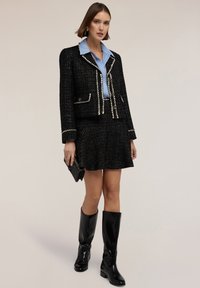 Donna che indossa un completo gonna giacca in tweed nero con bordi bianchi, camicia blu con colletto, tiene una borsa a tracolla nera e stivali neri fino al ginocchio, in piedi davanti a uno sfondo semplice.