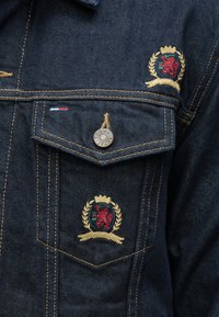 Σκούρο τζιν μπουφάν με χρυσοκεντήματα και έμβλημα με λιοντάρι. Διαθέτει αντίθετες ραφές και μεταλλικό κουμπί με λογότυπο της μάρκας.