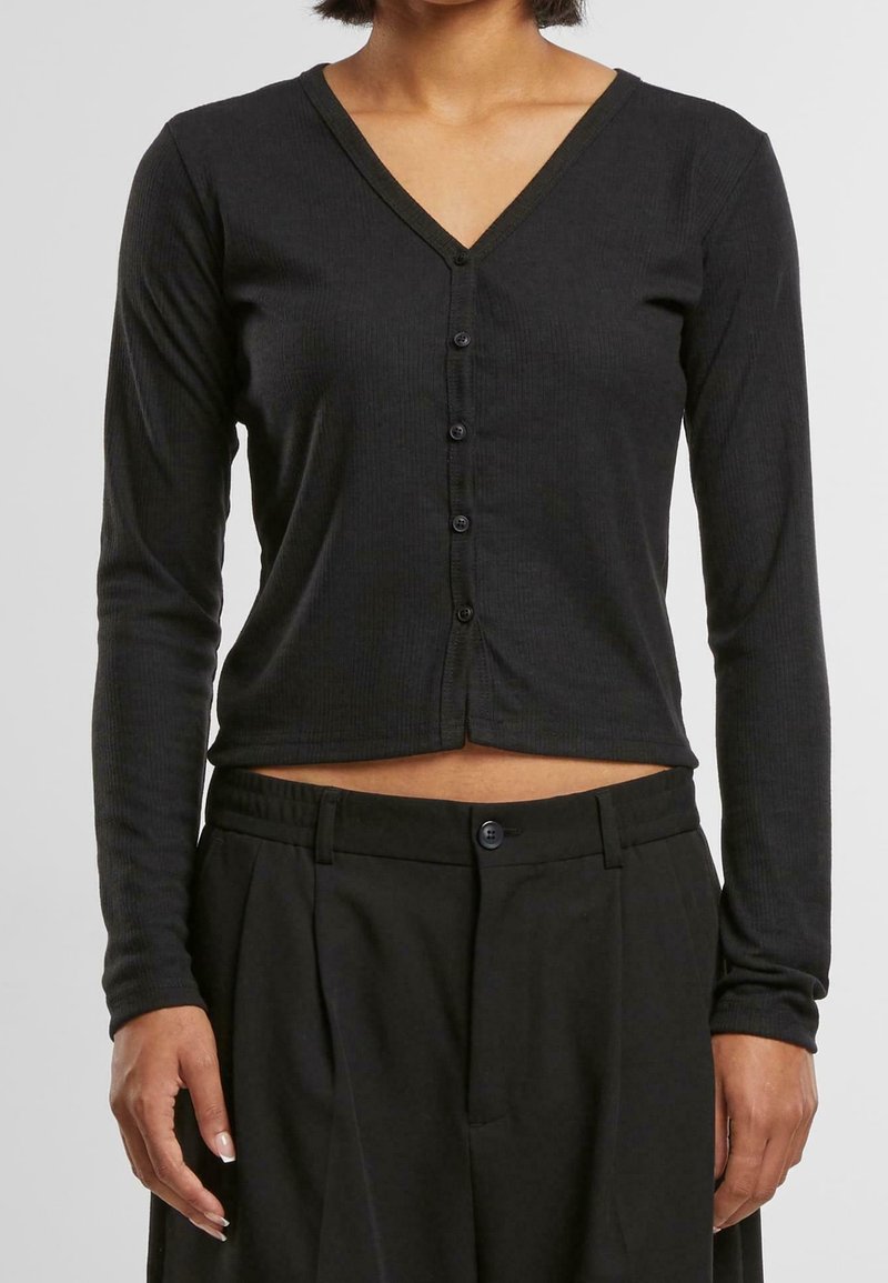 Femme portant un cardigan noir ajusté à manches longues, col en V et boutonné, ainsi qu’un pantalon noir plissé et ample sur un fond uni.