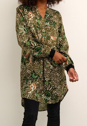 Femme portant une tunique vert olive avec des motifs floraux et abstraits multicolores, des poignets noirs et un pantalon noir, ajustant sa manche.