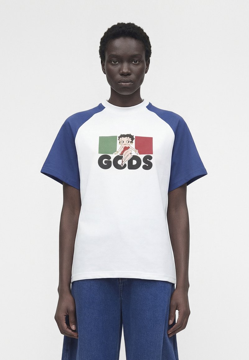 Άτομο που φοράει λευκό T-shirt με μπλε μανίκια, με χαρακτήρα κινουμένων σχεδίων και τα χρώματα της ιταλικής σημαίας πάνω από το έντονο κείμενο "GCDS", συνδυασμένο με μπλε παντελόνι.