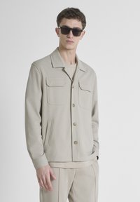 Lichtbeige jas met een kraag, twee borstzakken en een knoopsluiting aan de voorkant. Draagt over een bijpassend topje en in combinatie met broek in een vergelijkbare tint.