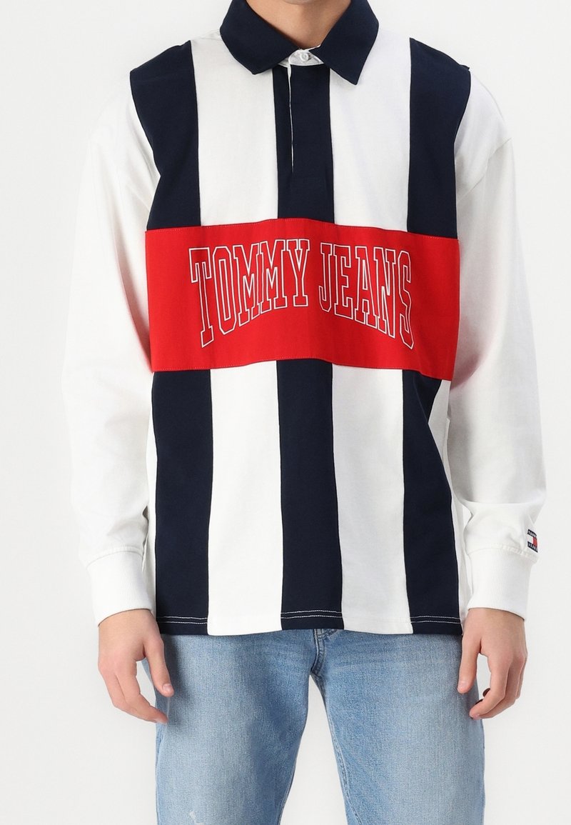 Long sleeve polo shirt in white, navy, and red. Features horizontal red band with "TOMMY JEANS" text. Made of cotton with a collared neckline.