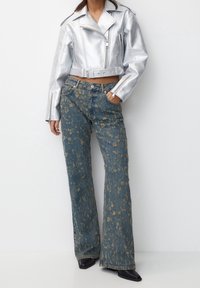 Veste de biker courte en argent métallique avec une taille ceinturée, associée à un jean flared bleu délavé orné de motifs floraux dorés et de détails usés.