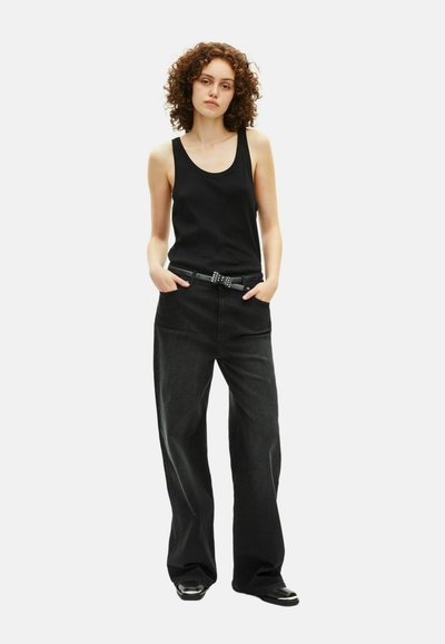 Top sin mangas negro y pantalones de pierna ancha gris oscuro con bolsillos frontales, cinturón negro texturizado con remaches decorativos y zapatos de suela plana.