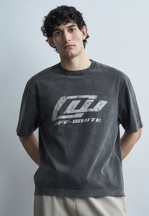 Jeune homme aux cheveux noirs bouclés portant un T-shirt graphique Off-White noir délavé et un pantalon de couleur claire, se tenant contre un fond uni.