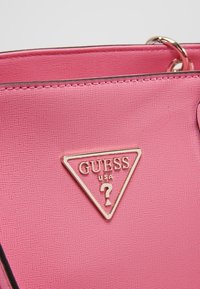 Sac à main synthétique rose avec finition texturée, doté d'une plaque logo triangulaire en or portant l'inscription "GUESS USA" et un design de point d'interrogation.