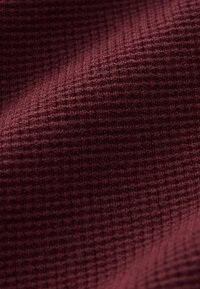Burgundy színű, textúrált szövet, ami gofriszerű szövési mintával rendelkezik, finom árnyalatváltozásokkal és puha, szőtt megjelenéssel.