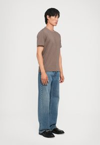 Brown patterned t-shirt with a crew neck, paired with loose-fit, light blue denim jeans and black shoes, seen from the side.