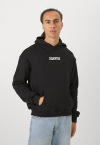 Čierna mikina z bavlneného zmesi, so zapínaním na prednej strane, rebrovanými manžetami a bielym logom "JUVENTUS" na hrudi. V kombinácii so svetlými džínsami.