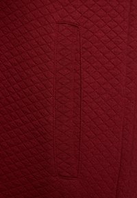 Tissu matelassé bourgogne avec un motif en losanges et un détail de poche verticale. La texture semble douce et densément tissée.