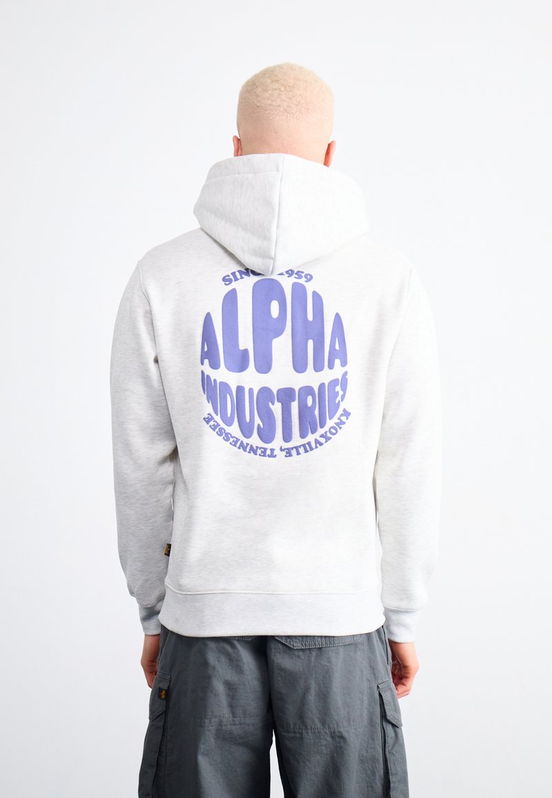 Šviesiai pilkas ilgomis rankovėmis džemperis su dideliu violetiniu apskritu logotipu, rašančiu "ALPHA INDUSTRIES", ant nugaros. Turi kapšą su raišteliu ir įgulos sandarumo rankoves.