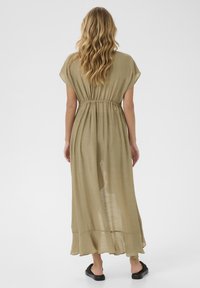 Long, olive green dress made from lightweight fabric. Features short sleeves, an elastic waist, and a flowing hemline. Flat sandals complete the look.