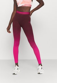 Kobieta ubrana w legginsy w gradient od fioletowego do różowego, różowy stanik sportowy i białe buty sportowe, stoi z jedną ręką na biodrze na tle gładkiej ściany.