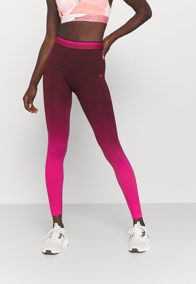 Kobieta ubrana w legginsy w gradient od fioletowego do różowego, różowy stanik sportowy i białe buty sportowe, stoi z jedną ręką na biodrze na tle gładkiej ściany.