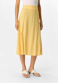 Jupe midi jaune à pois blancs, avec une fermeture sur le côté par bouton, en tissu lisse et une silhouette évasée. Sandales à semelle plate visibles.