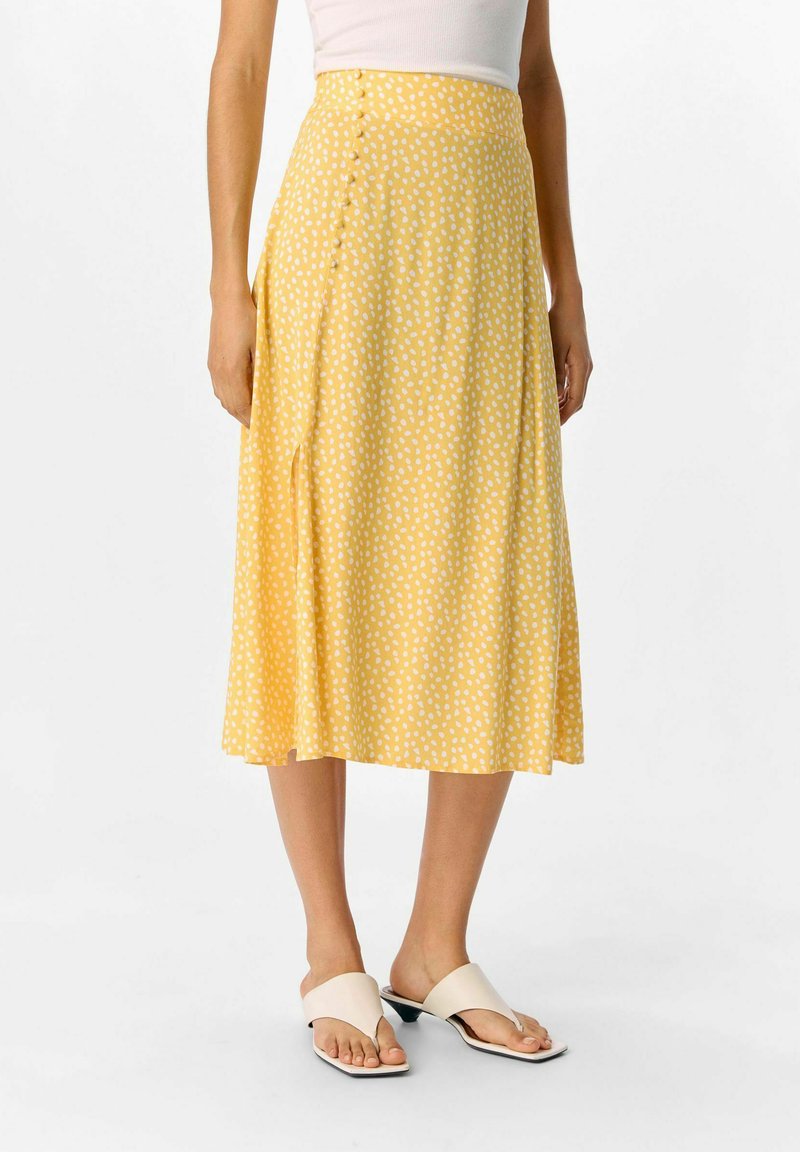 Jupe midi jaune à pois blancs, avec une fermeture sur le côté par bouton, en tissu lisse et une silhouette évasée. Sandales à semelle plate visibles.