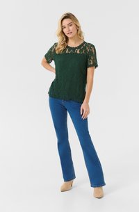Top verde in pizzo con maniche corte, scollatura rotonda e orlo a onda. Abbinato a jeans blu a palloncino e stivaletti beige con tacco. Sfondo bianco semplice.