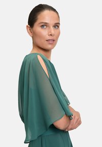 Abito in chiffon verde con maniche corte e design con spalle scoperte, caratterizzato da una silhouette fluida e una texture liscia. Modella posata contro uno sfondo bianco.