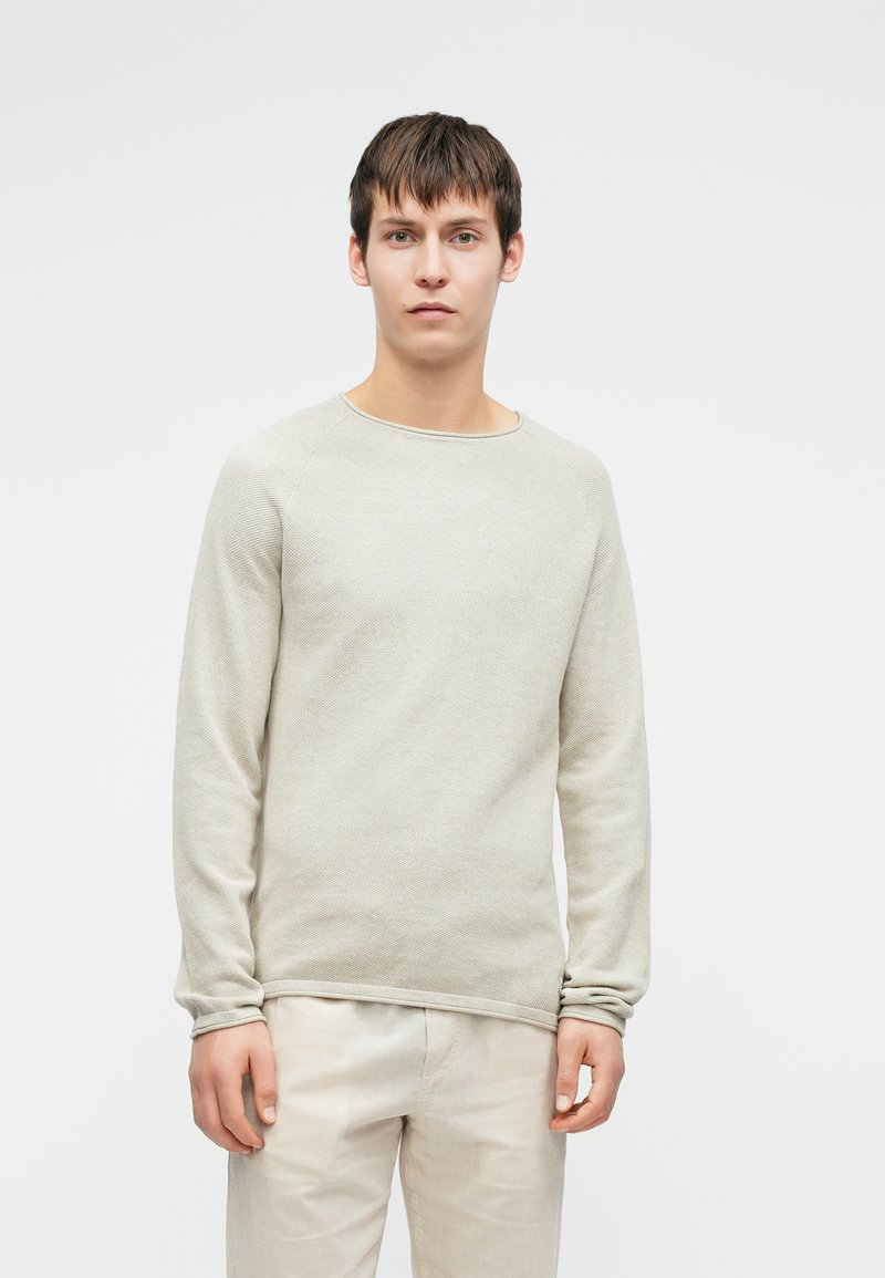Maglione grigio chiaro a maniche lunghe realizzato in tessuto a maglia testurizzata, caratterizzato da un ampio scollo rotondo e polsini a costine. Indossato con pantaloni beige chiaro.