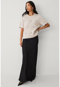 Beiger Strickpullover mit kurzen Ärmeln, verziert mit silbernen Blumendetails, kombiniert mit einem schwarzen Satin-Maxirock und spitzen schwarzen Schuhen.