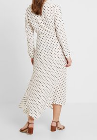 Långärmad, krämfärgad klänning med bruna prickar, utsvängd nederkant och en omslagdesign. Har en sidoklyvning och är matchad med bruna klacksandaler.