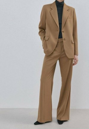 Mujer con traje a medida color beige con pantalones de pierna ancha y un cuello vuelto negro, de pie con una mano en el bolsillo contra un fondo liso.