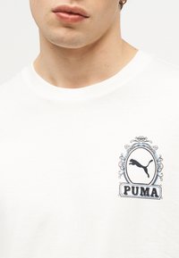 Κοντινό πλάνο ενός λευκού t-shirt με διακοσμητικό λογότυπο Puma σε μαύρο και μπλε στην αριστερή πλευρά του στήθους, που φοριέται από ένα άτομο με κρίκο χειλιού και σκουλαρίκι.