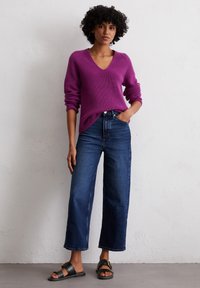 Lila gerippter Pullover mit V-Ausschnitt, kombiniert mit hochtaillierten, dunkelblauen Schlagjeans und schwarzen Sandalen mit einer goldenen Schnalle.