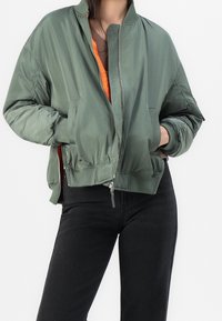 Personne portant une veste bomber verte surdimensionnée avec une doublure orange, pantalon noir, mains partiellement dans les poches de la veste, debout devant un fond blanc.