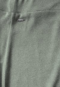 Grüner Strickstoff mit einer strukturierten Oberfläche und einem silbernen Logo-Tag, das darauf genäht ist. Das Material wirkt weich und flexibel.