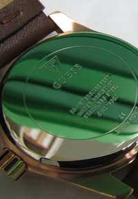 Round stainless steel watch back with green reflective surface, engraved with brand name "Guess," water resistance info, and model details.