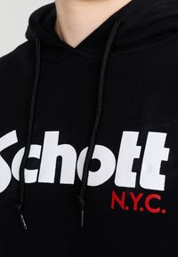 Felpa nera con cappuccio caratterizzata da un grande logo bianco "Schott" e il testo rosso "N.Y.C.". Presenta cordini con estremità in metallo e una texture morbida.
