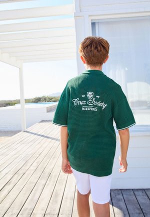 Rövid barna hajú gyermek zöld, struktúrált pólót visel, amelyen a "Cruz Society Palm Springs" felirat olvasható a hátoldalán, és egy napfényes faerkélyen sétál, közel egy fehér épülethez.