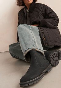 Schwarze Knöchelstiefel mit Gummisohlen und elastischen Seitenpaneelen, kombiniert mit hellblauen Jeans und einer gesteppten schwarzen Jacke, auf neutralem Hintergrund.