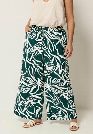 Kvinde iført grønne bukser med vide ben og hvidt blomsterprint, kombineret med en beige ærmeløs top og hvide slid-sandaler.