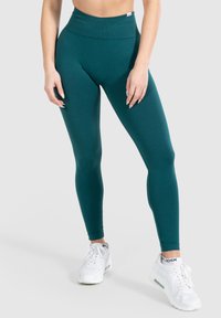 Slim fit teal leggings gedragen door een staand persoon, gecombineerd met witte sportschoenen op een lichte achtergrond.