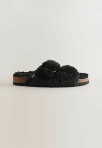 Sorte fluffy slip-on slippers med to pom-pom accenter, kork mellemsål og en tekstureret sort ydersål for bedre greb. Blød faux fur overflade.