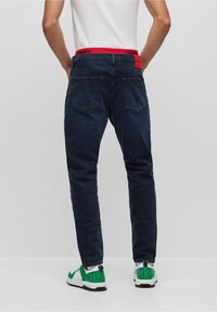 Jeans de mezclilla azul oscuro con corte recto, que cuentan con una cinturilla roja de HUGO y bolsillos traseros. Se llevan con zapatillas verdes y blancas.