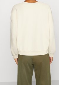 Sweat-shirt oversize de couleur crème, à col rond, épaules tombantes et poignets côtelés. Le tissu semble doux et lisse.