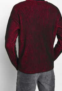 Pull rayé noir et rouge à manches longues, col rond, et tricot texturé. Le design présente des rayures verticales et une coupe décontractée.