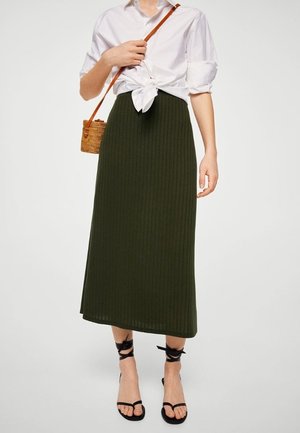 Femme portant une chemise blanche nouée, une jupe midi côtelée vert foncé, des sandales noires à lacets à la cheville, et portant un petit sac bandoulière tissé.