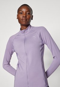 Violet ilgos rankovės sportinė zip-up striukė su briaunotu tekstūru, turinti aukštą apykaklę ir mažą atspindinčią logotipo detalę ant krūtinės.