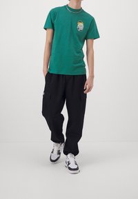 Vihreä lyhythihainen t-paita, jossa rintakuva, yhdistettynä löysiin mustiin cargo-housuihin ja valkoisiin lenkkareihin mustilla yksityiskohdilla.