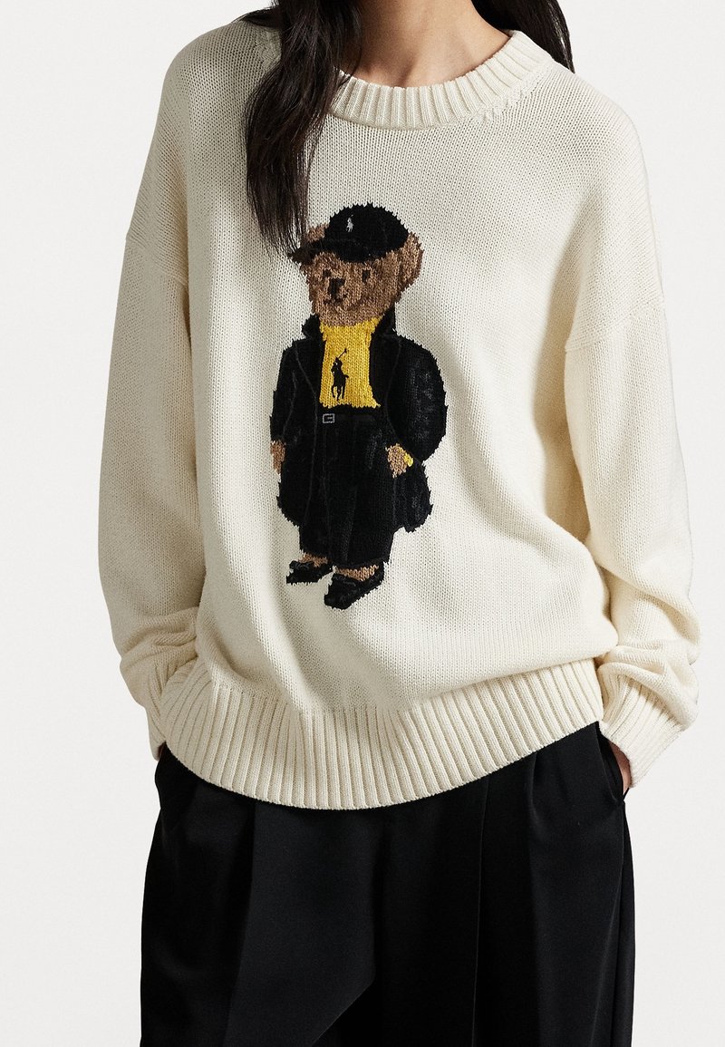 Pull en maille crème avec un ours en peluche portant un manteau noir, une chemise jaune et une casquette noire, porté par une personne aux cheveux foncés et un pantalon noir.
