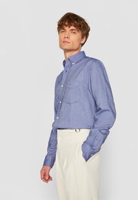 Langärmliges Chambray-Hemd in Hellblau mit Button-Down-Kragen, Brusttasche und gerader Passform, kombiniert mit hochtaillierten beigen Hosen.