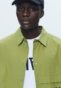 Green button-up shirt with a structured collar, paired with a white graphic t-shirt. The shirt features snap fasteners and a smooth texture.