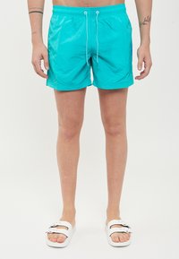 Pantaloncini da bagno turchesi realizzati in materiale leggero, dotati di una vita elasticizzata con cordoncino e tasche laterali. Indossati con sandali bianchi.