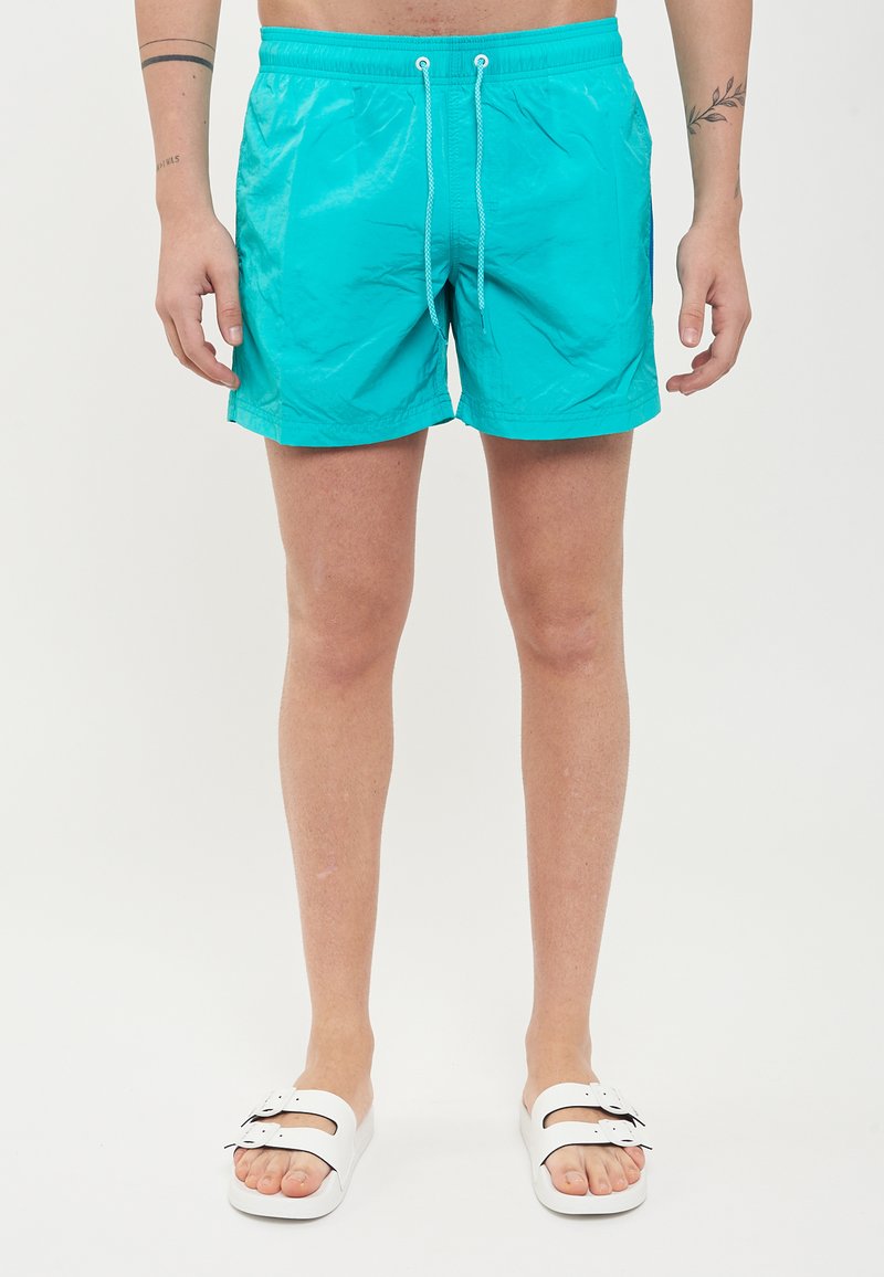 Pantaloncini da bagno turchesi realizzati in materiale leggero, dotati di una vita elasticizzata con cordoncino e tasche laterali. Indossati con sandali bianchi.