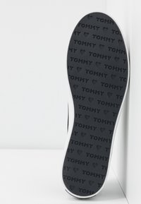 Semelle de chaussure noire en caoutchouc présentant un motif texturé avec les inscriptions "TOMMY" et des motifs de cœur, bordée d'un contour blanc, sur un fond clair.